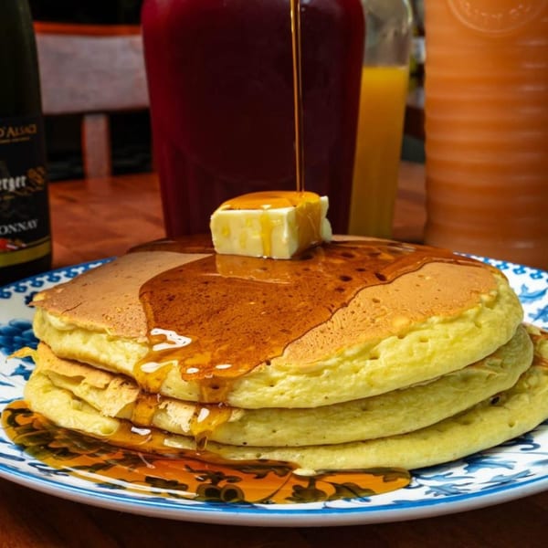 Maple syrup pouring over a stack of golden pancakes with butter on a blue and white plate.