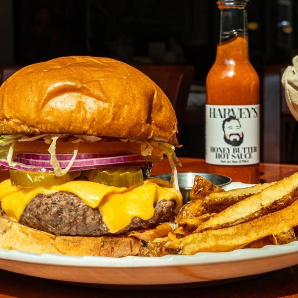 A Harvey's cheeseburger with fries and a bottle of Honey Butter Hot Sauce on a white plate.